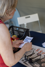 Kobieta w czerwonej bluzce trzyma w dłoniach złożoną ulotkę z napisem „SENIORZE! BĄDŹ AKTYWNY”. W tle na stole leżą gazety, okulary przeciwsłoneczne oraz kosz z jabłkami.