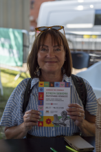 Portret uśmiechniętej pracowniczki Biura Trzeciego Wieku w okularach przeciwsłonecznych na głowie i paskowanej bluzce, prezentującej do kamery ulotkę „Strefa Seniora Przystanek Równość”.