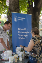 Mateusz Baluk, prowadzący warsztaty stoi przy stoisku z niebieskim roll-upem "Uniwersytet Gdański – Gdański Uniwersytet Trzeciego Wieku" rozmawia z uczestniczką plenerowych warsztatów kosmetycznych.